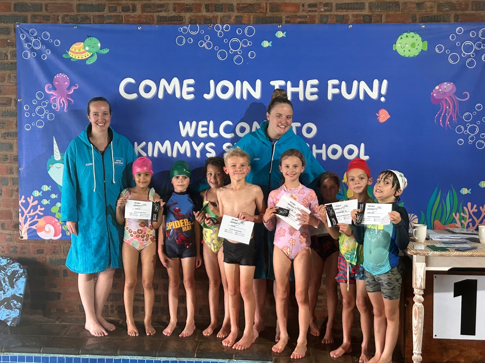 Swimmers with certificates in front of the Kimmy's Swim School banner